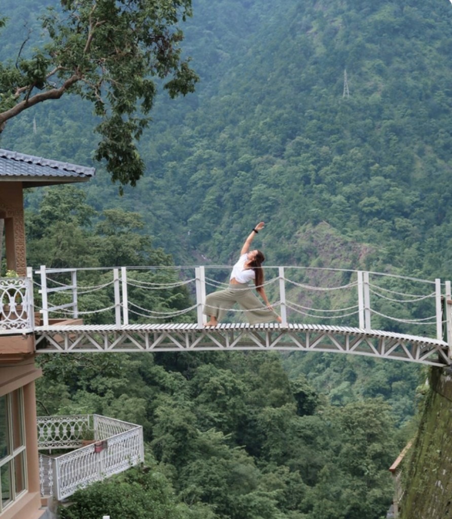 Júlia Sans on a footbridge above a forested valley, one arm lifted, hair down her back.